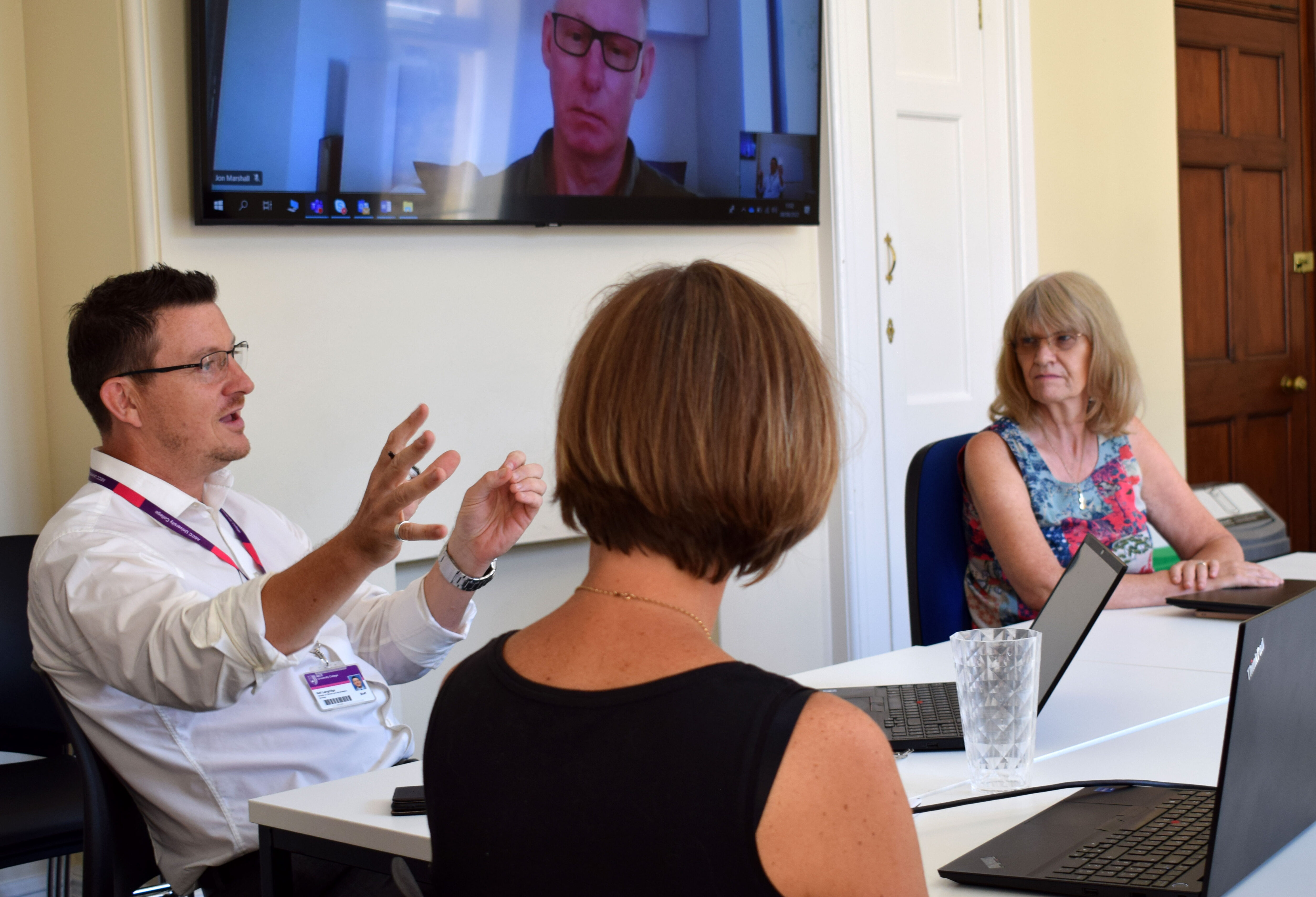 clinicians in a hybrid meeting with male colleague calling in remotely via a large screen and four colleagues in a meeting room