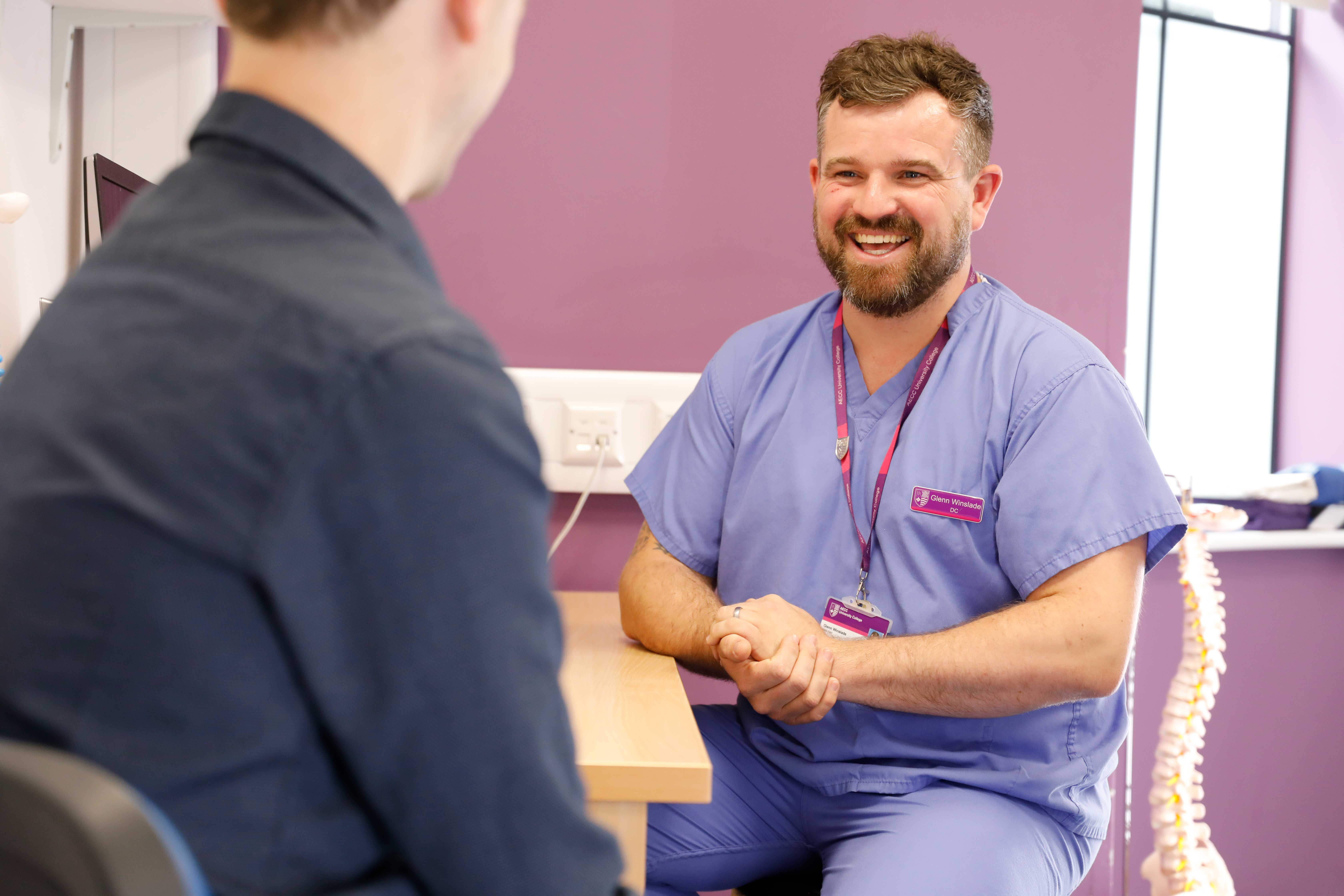 Over the shoulder shot of male chiropractor in consultation with patient. The chiropractor has a jolly smile and clasps his hands together in a warm friendly manner